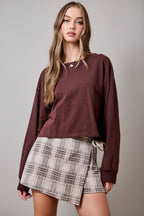 A woman wearing an oatmeal brown and cream plaid wrap skort with a side tie, paired with brown sweater set against a solid tan studio background.