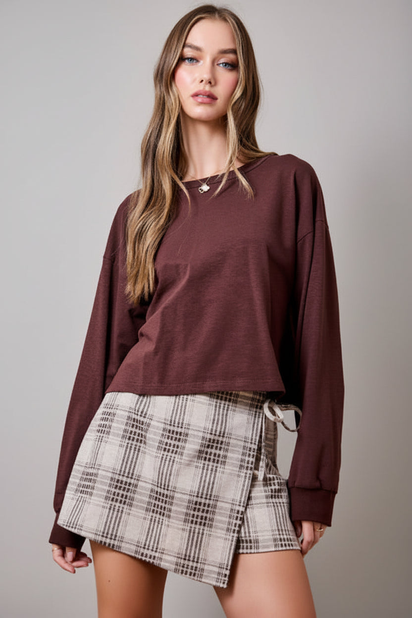A woman wearing an oatmeal brown and cream plaid wrap skort with a side tie, paired with brown sweater set against a solid tan studio background.