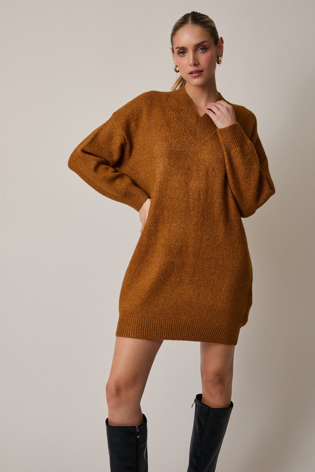 Woman wearing a camel sweater dress with black knee-high boots on a plain background