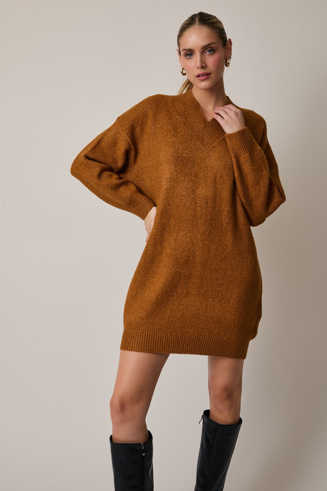 Woman wearing a camel sweater dress with black knee-high boots on a plain background