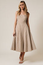 A woman wearing a sleeveless midi dress with thin vertical stripes in beige and brown, photographed from the back against a solid warm beige studio backdrop.