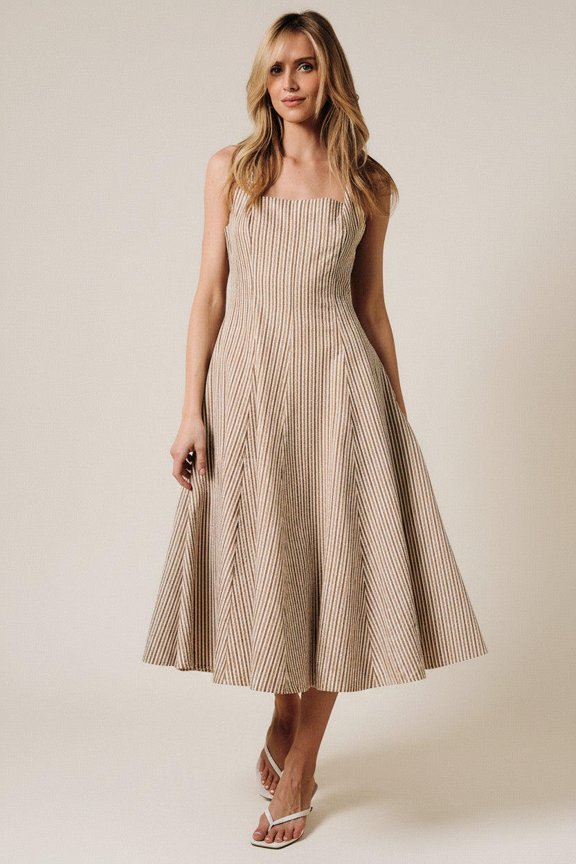 A woman wearing a sleeveless midi dress with thin vertical stripes in beige and brown, photographed from the back against a solid warm beige studio backdrop.