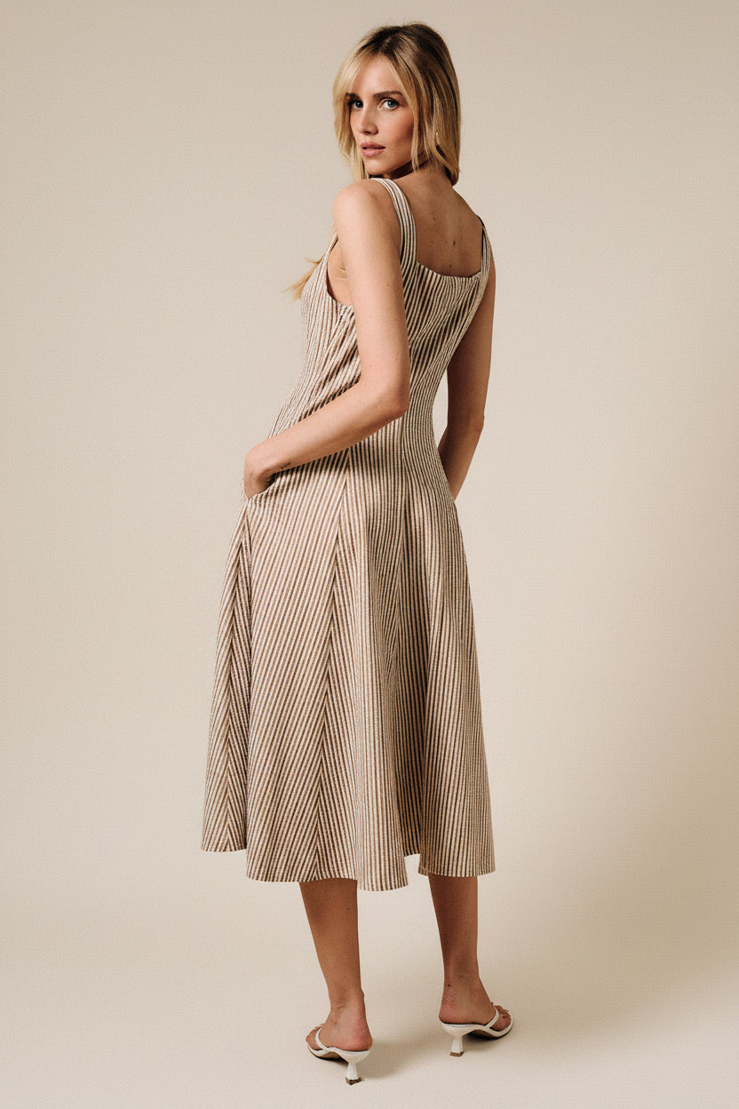 A woman wearing a sleeveless midi dress with thin vertical stripes in beige and brown, photographed from the back against a solid warm beige studio backdrop.