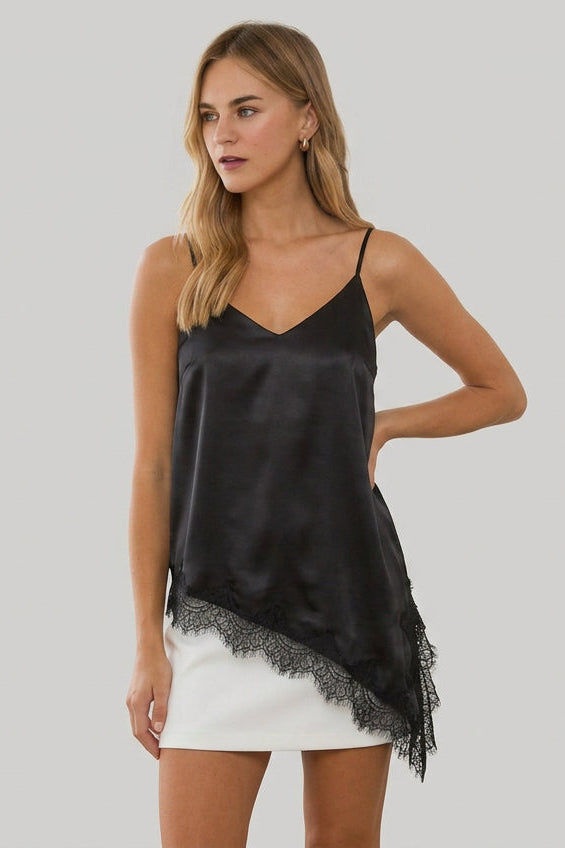 A woman modeling a black spaghetti-strap crinkled satin cami tunic with lace hem and a white leather mini skirt, standing in a bright, sunlit room with white columns.