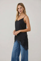 A woman modeling a black spaghetti-strap crinkled satin cami tunic with lace hem and a black faux-leather mini skirt, standing in a bright, sunlit room with white columns.