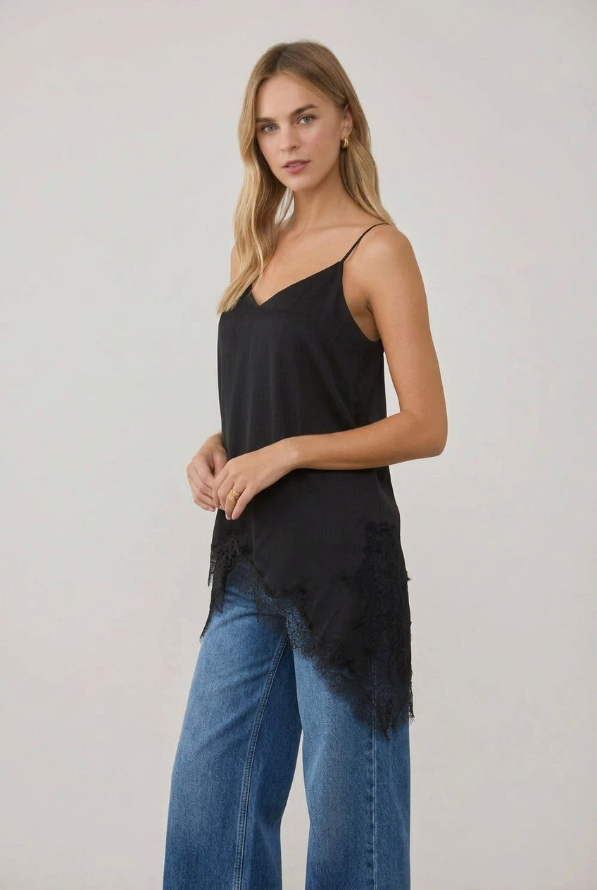 A woman modeling a black spaghetti-strap crinkled satin cami tunic with lace hem and a black faux-leather mini skirt, standing in a bright, sunlit room with white columns.
