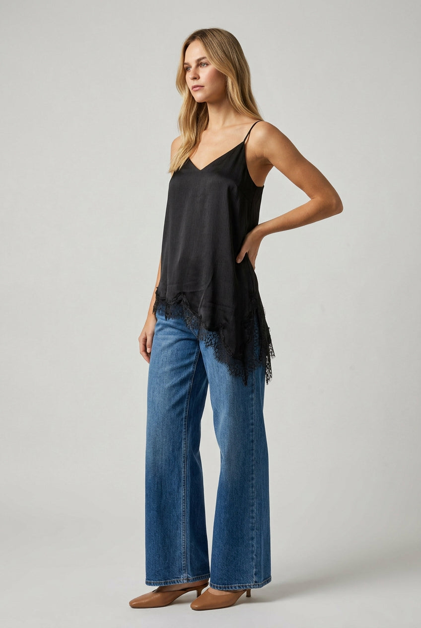 A woman modeling a black spaghetti-strap crinkled satin cami tunic with lace hem and jeans standing in a bright, sunlit room with white columns.