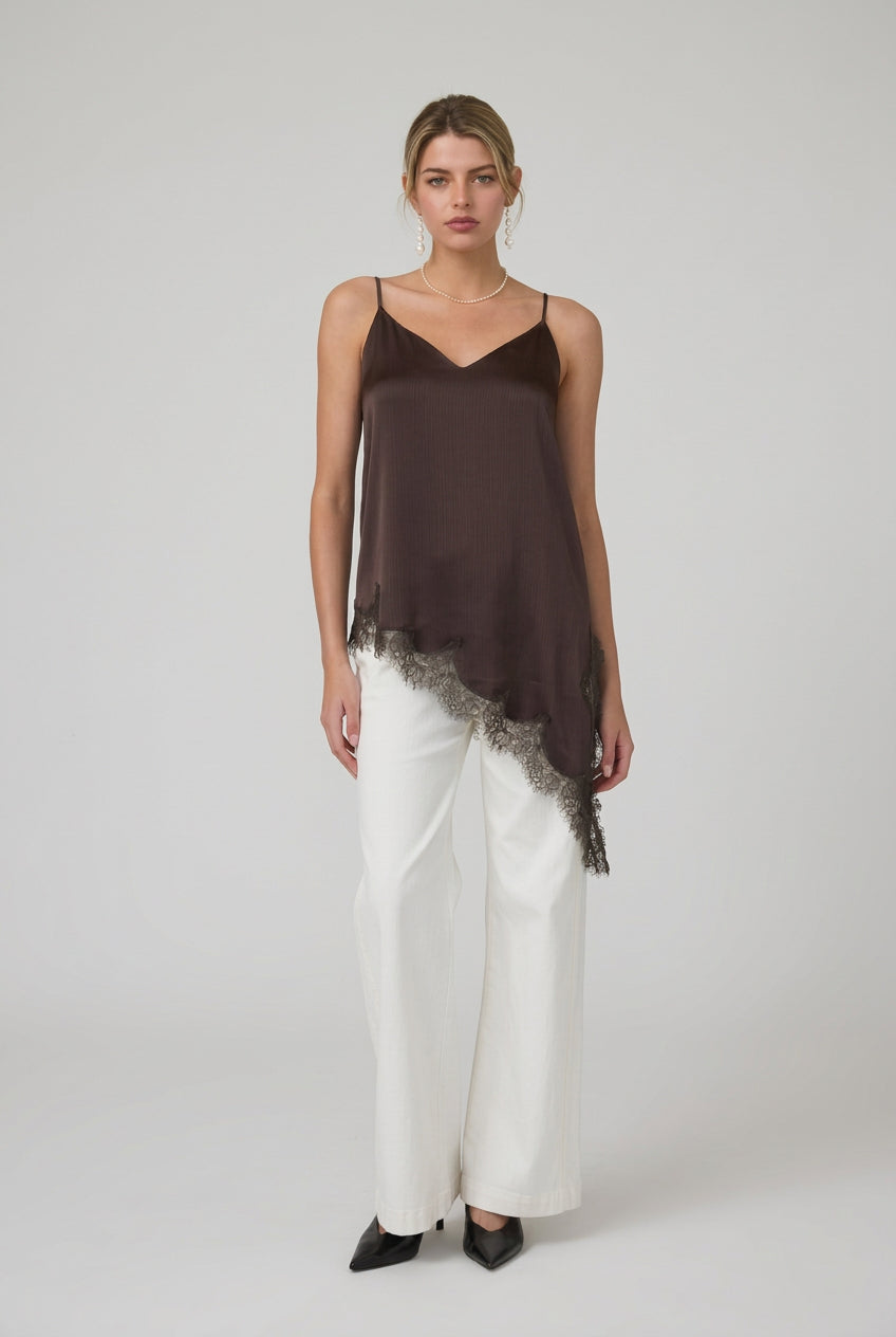 Woman wearing a brown satin asymmetrical lace hemmed cami tunic top and white pants in a minimalistic room.