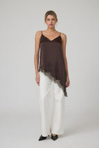 Woman wearing a brown satin asymmetrical lace hemmed cami tunic top and white pants in a minimalistic room.