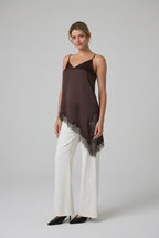 Woman wearing a brown satin asymmetrical lace hemmed cami tunic top and white pants in a minimalistic room.