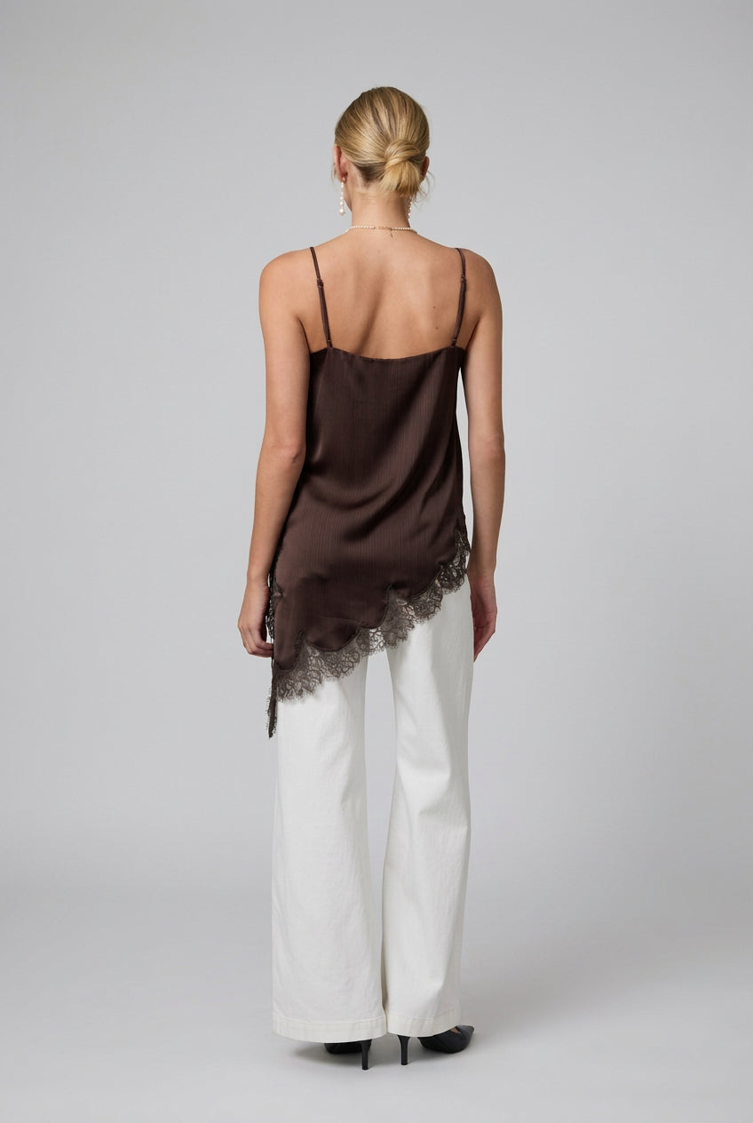 Back view of a woman wearing a brown satin asymmetrical lace hemmed cami tunic top and white pants in a minimalistic room.