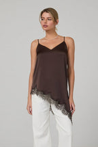 Woman wearing a brown satin asymmetrical lace hemmed cami tunic top and white pants in a minimalistic room.