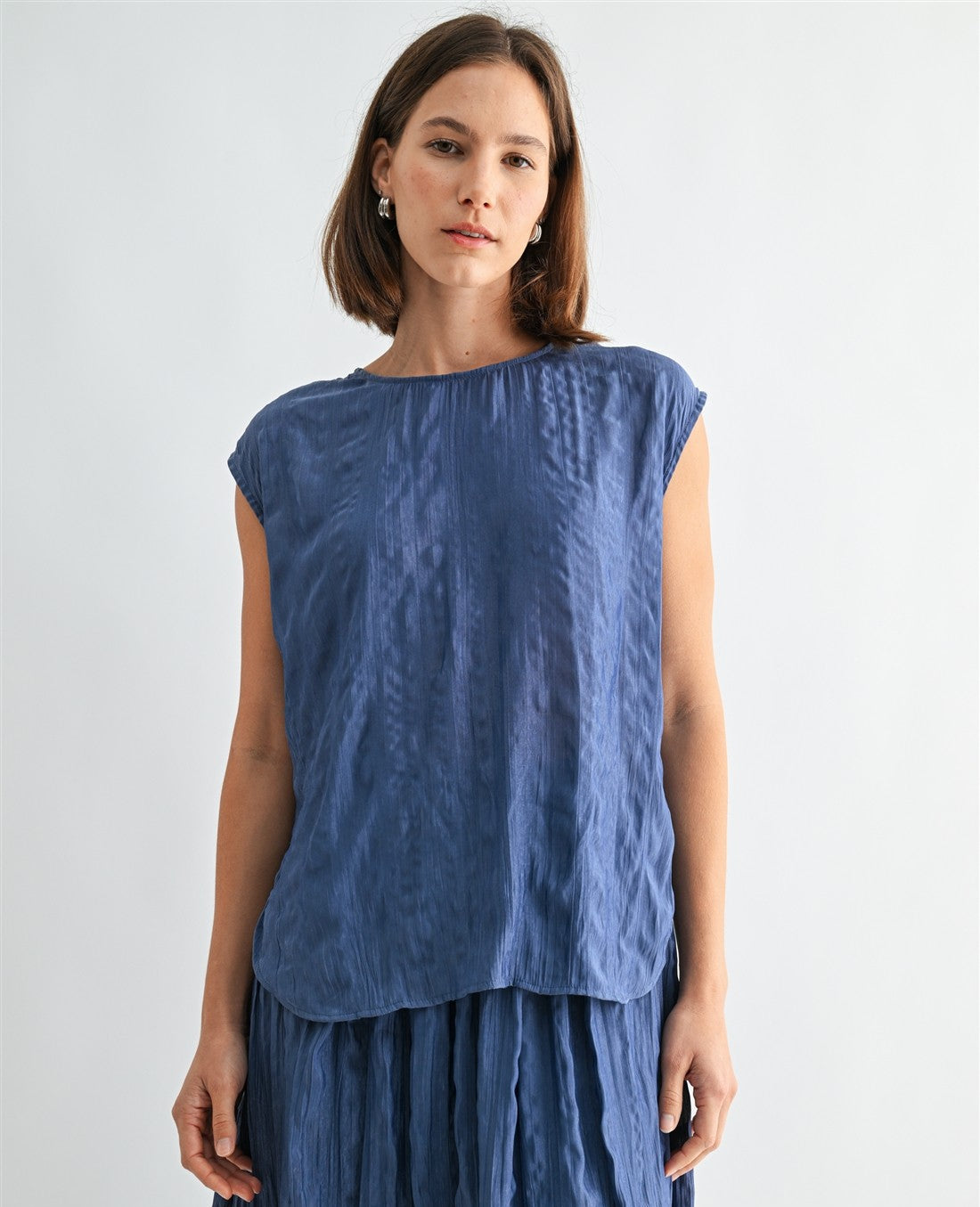 Woman wearing a dusty blue crinkle satin cap sleeve top against a light gray background