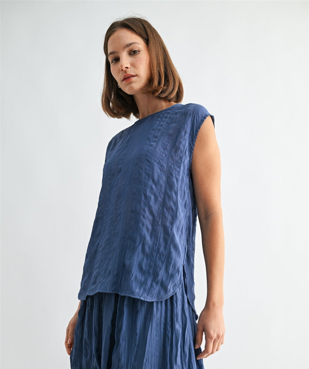 Woman wearing a dusty blue crinkle satin cap sleeve top against a light gray background