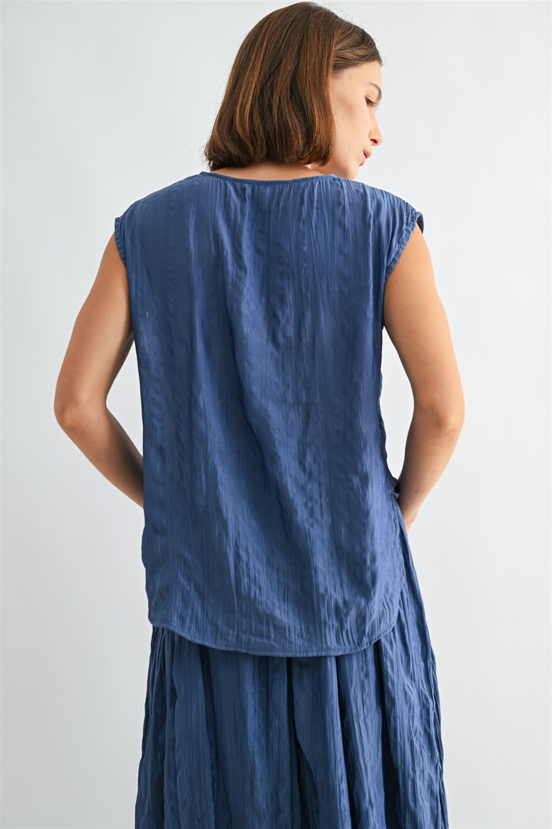Back view of a woman wearing a dusty blue crinkle satin cap sleeve top against a light gray background
