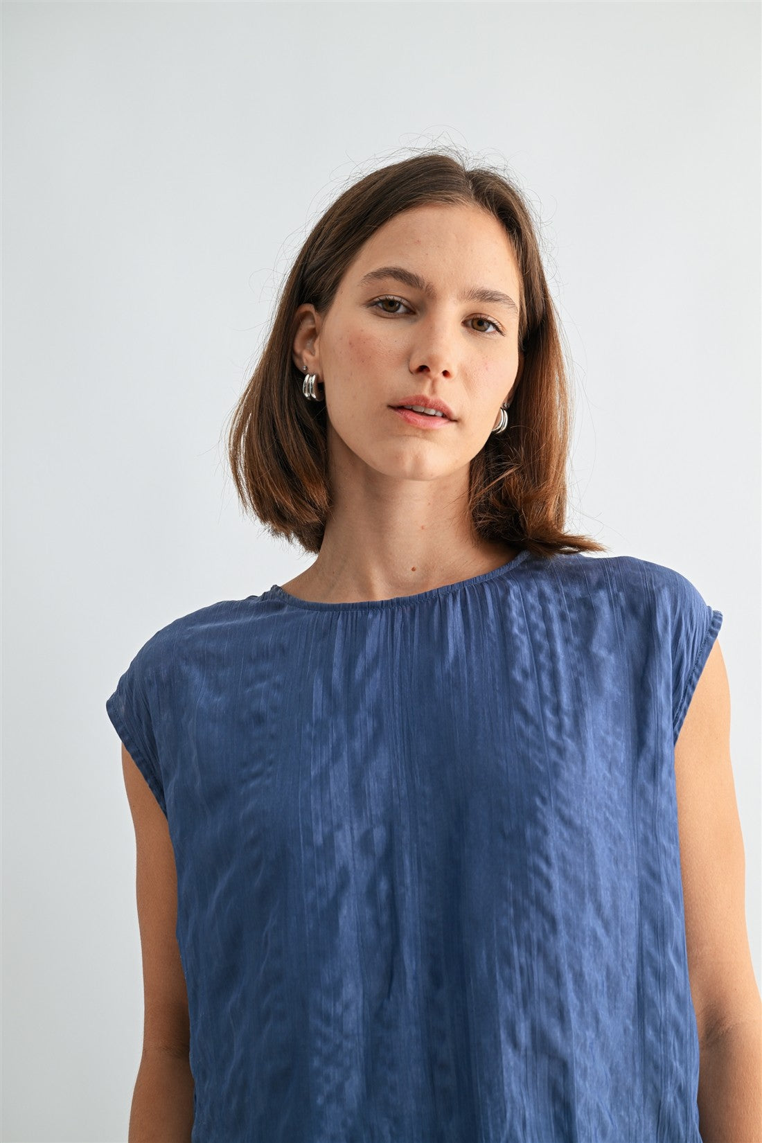 Woman wearing a dusty blue crinkle satin cap sleeve top against a light gray background