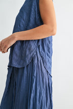 Side view of a woman wearing a dusty blue crinkle satin cap sleeve top against a light gray background