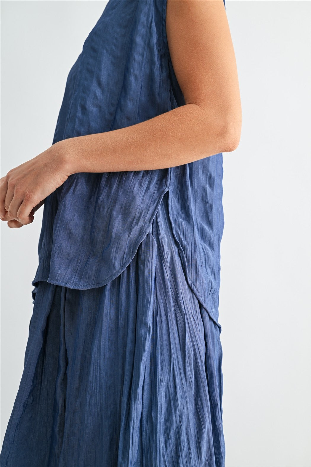 Side view of a woman wearing a dusty blue crinkle satin cap sleeve top against a light gray background