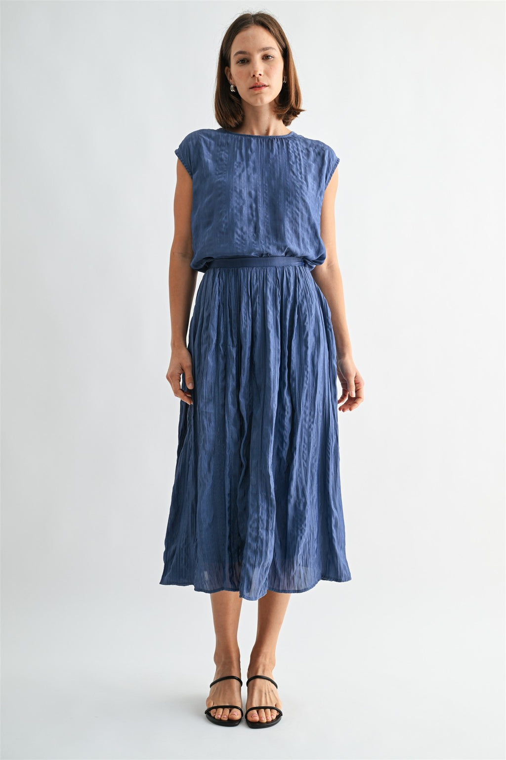 Woman wearing a dusty blue crinkle satin cap sleeve top and skirt against a light gray background