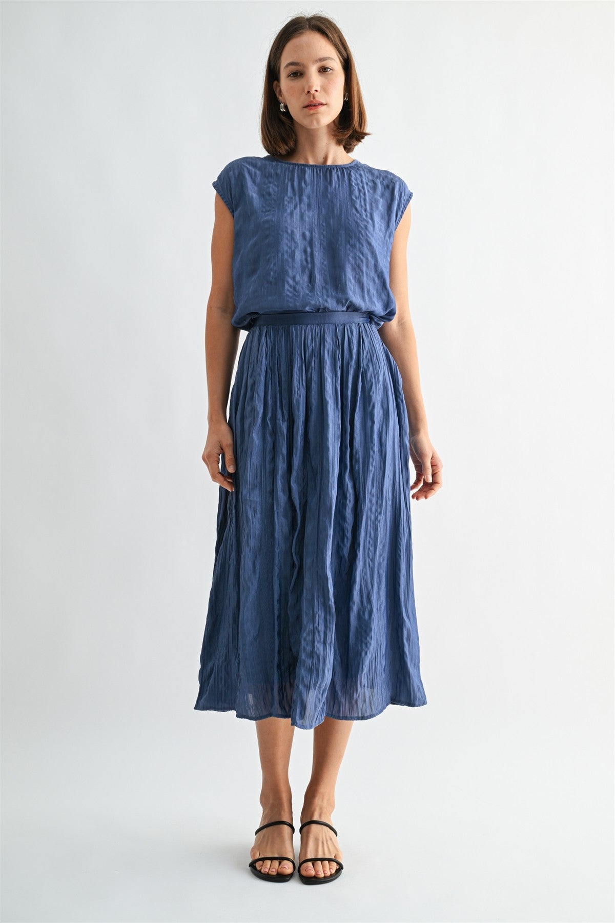 Woman wearing a dusty blue crinkle satin cap sleeve top and skirt against a light gray background