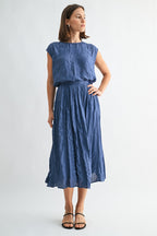 Woman wearing a dusty blue crinkle satin cap sleeve top and skirt against a light gray background