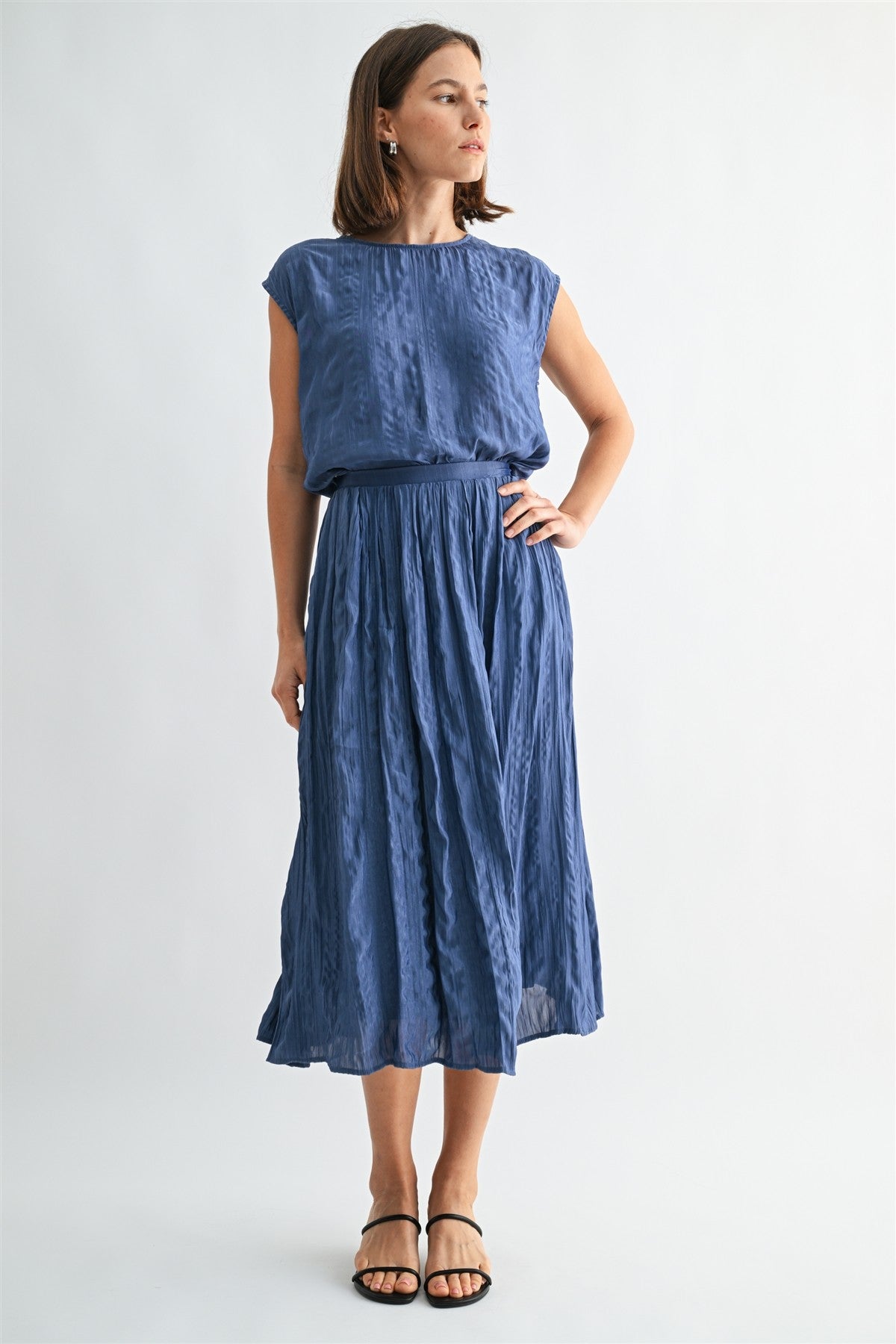 Woman wearing a dusty blue crinkle satin cap sleeve top and skirt against a light gray background