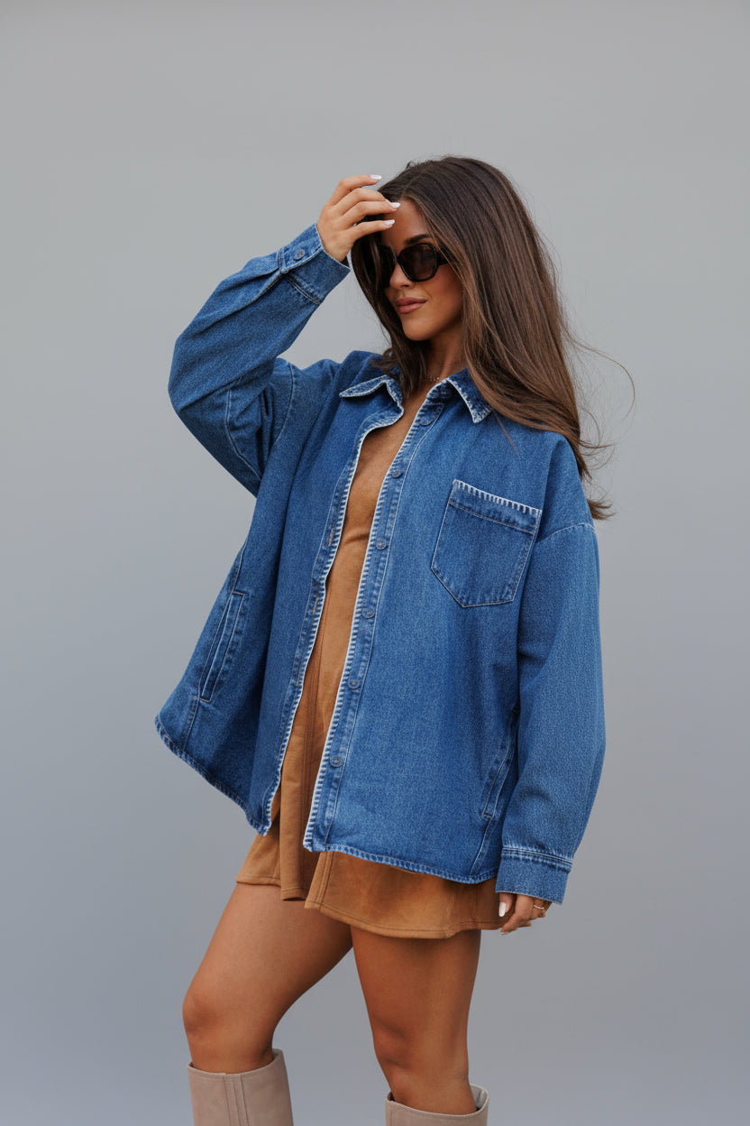 Woman wearing a blue denim shacket with contrast stitching over a brown dress against a stone wall.