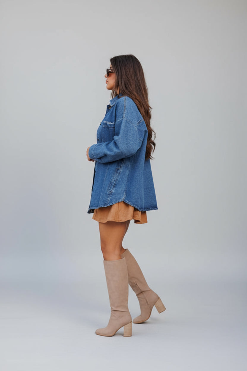Woman wearing a blue denim shacket with contrast stitching over a brown dress against a stone wall.