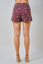 Back view of a woman wearing maroon and white floral embroidered eyelet shorts with side pockets, shown against a light beige background.