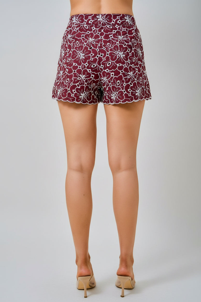 Back view of a woman wearing maroon and white floral embroidered eyelet shorts with side pockets, shown against a light beige background.