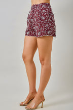A woman wearing maroon and white floral embroidered eyelet shorts with side pockets, shown against a light beige background.