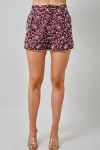 A woman wearing maroon and white floral embroidered eyelet shorts with side pockets, shown against a light beige background.