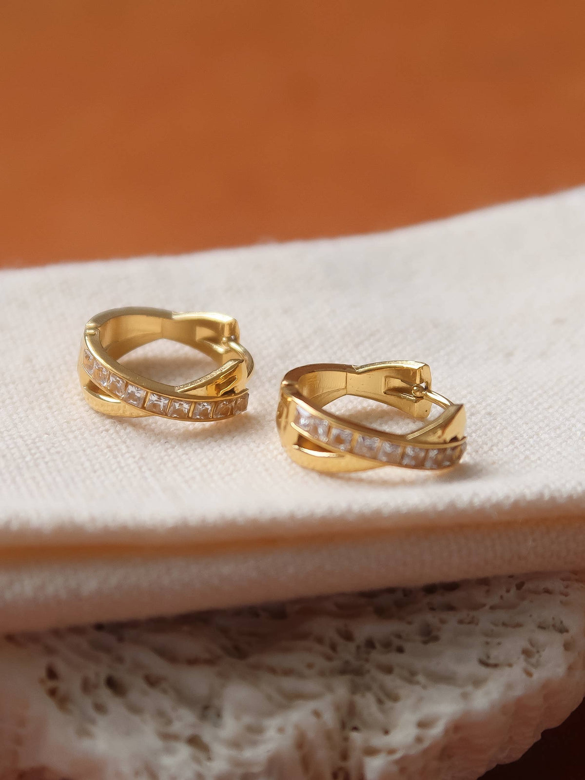 A pair of gold huggie hoop earrings with pavé-set clear stones, resting on a textured cream fabric surface with a warm brown background.