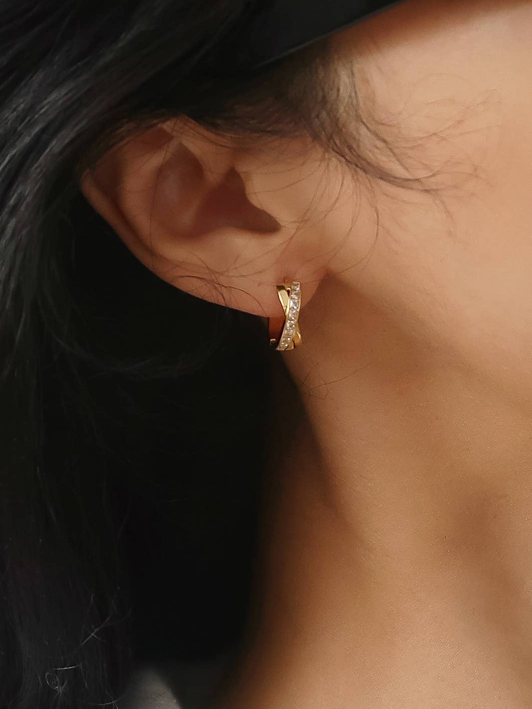 A close-up of a person's ear wearing a small gold huggie hoop earring with clear stones, against dark hair and warm skin tones.