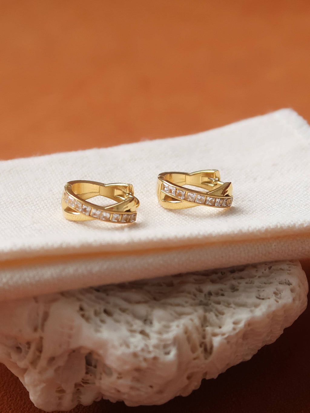 A pair of gold huggie hoop earrings with pavé-set clear stones, displayed on a folded cream fabric surface with a textured stone prop against a warm brown background.