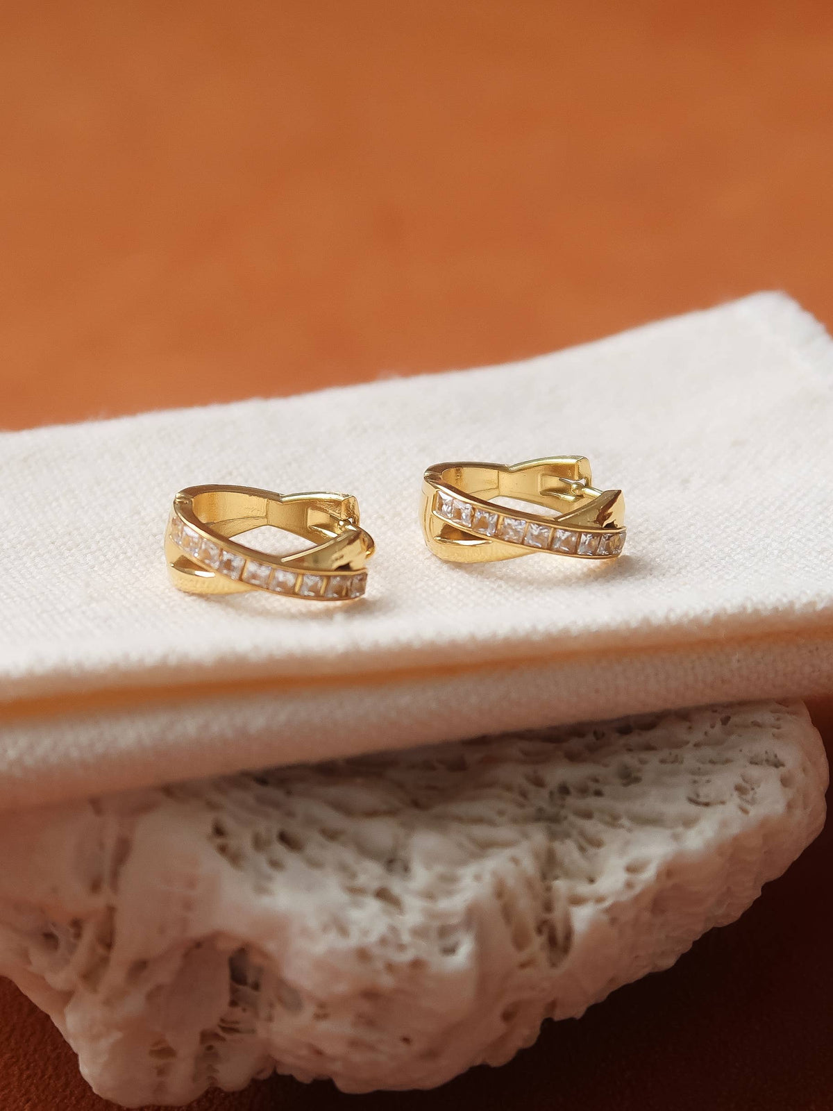 A pair of gold huggie hoop earrings with pavé-set clear stones, displayed on a folded cream fabric surface with a textured stone prop against a warm brown background.