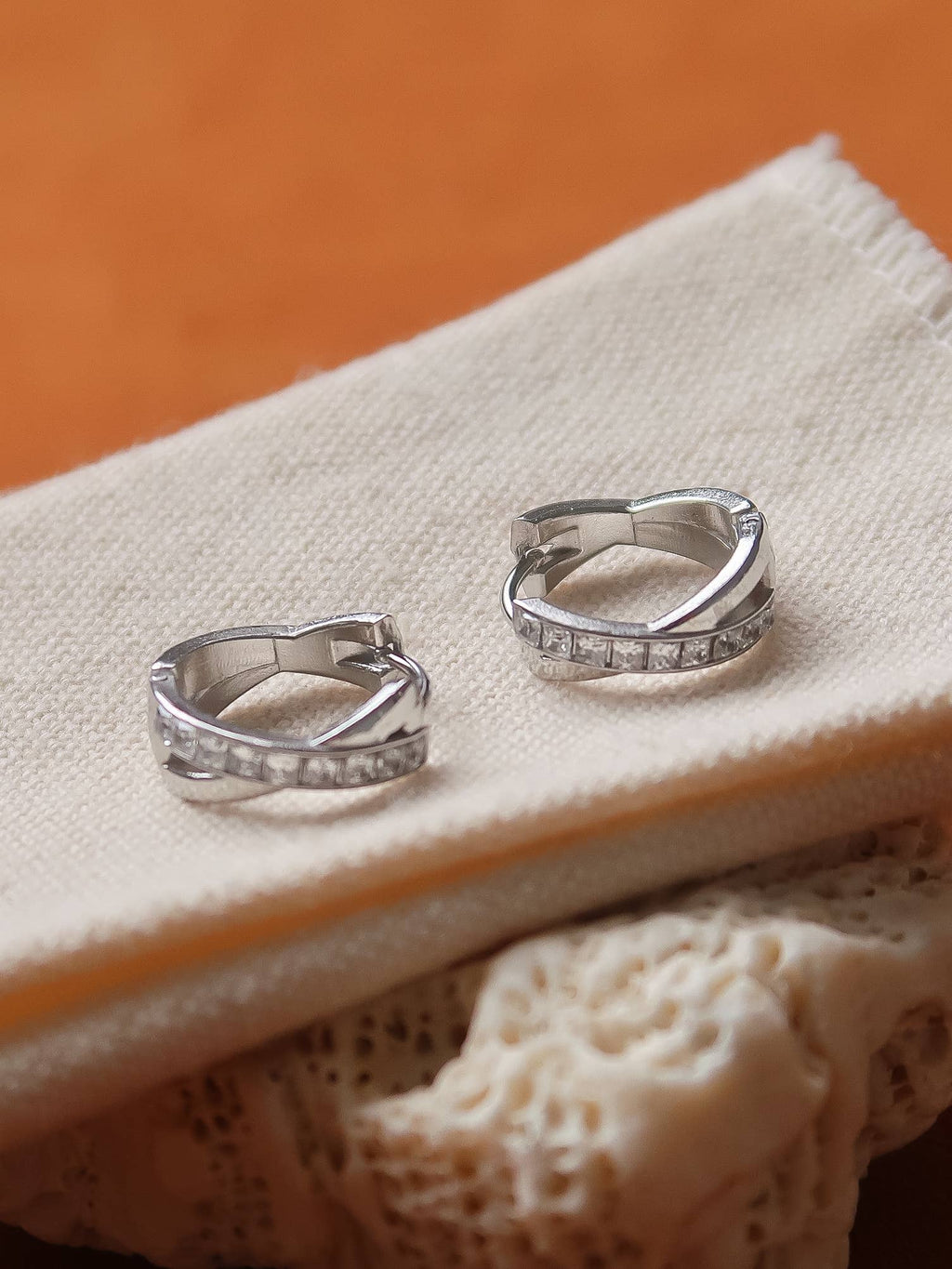 A pair of silver huggie hoop earrings with small CZ stones, resting on a textured cream fabric over a lace surface with a warm orange background.