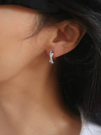 A close-up of a person's ear wearing a small silver huggie hoop earring with clear stones.