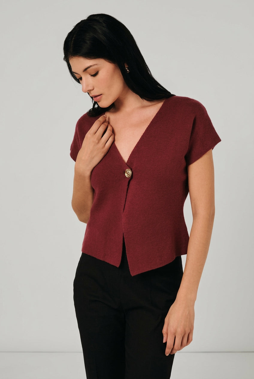 A woman wearing a short-sleeve burgundy knit top with a single decorative button, asymmetrical hemline and black pants, standing against a light curtain backdrop.