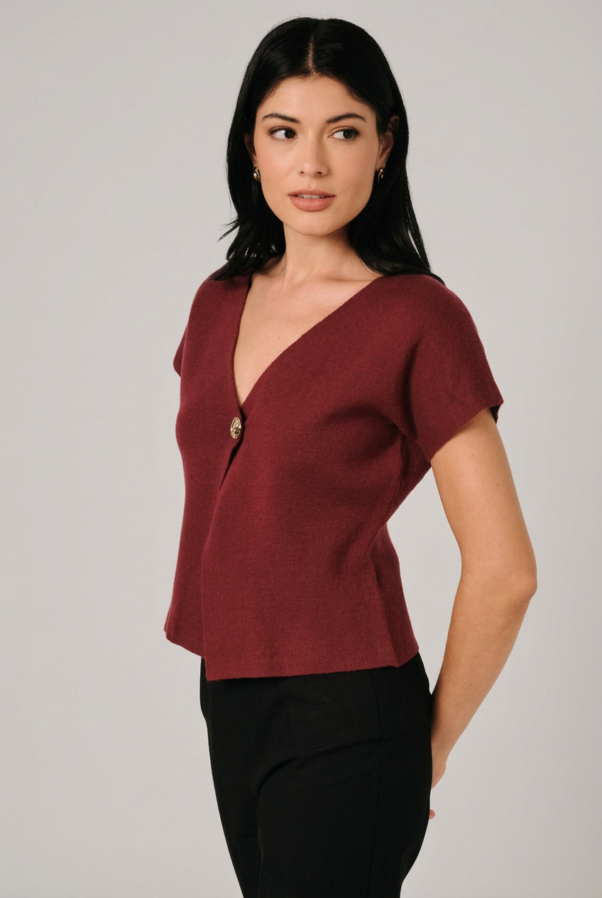 A woman wearing a short-sleeve burgundy knit top with a single decorative button, asymmetrical hemline and black pants, standing against a light curtain backdrop.