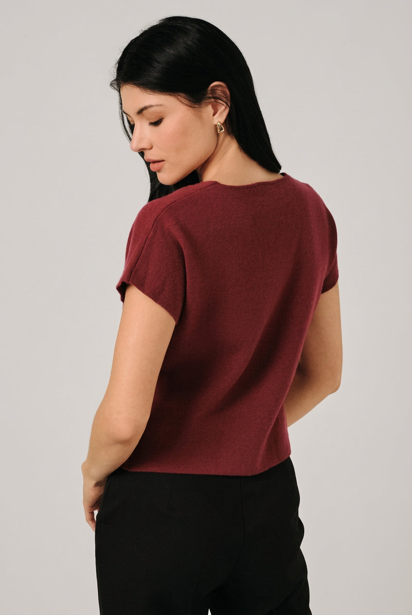 Back view of a woman wearing a short-sleeve burgundy knit top with a single decorative button, asymmetrical hemline and black pants, standing against a light curtain backdrop.