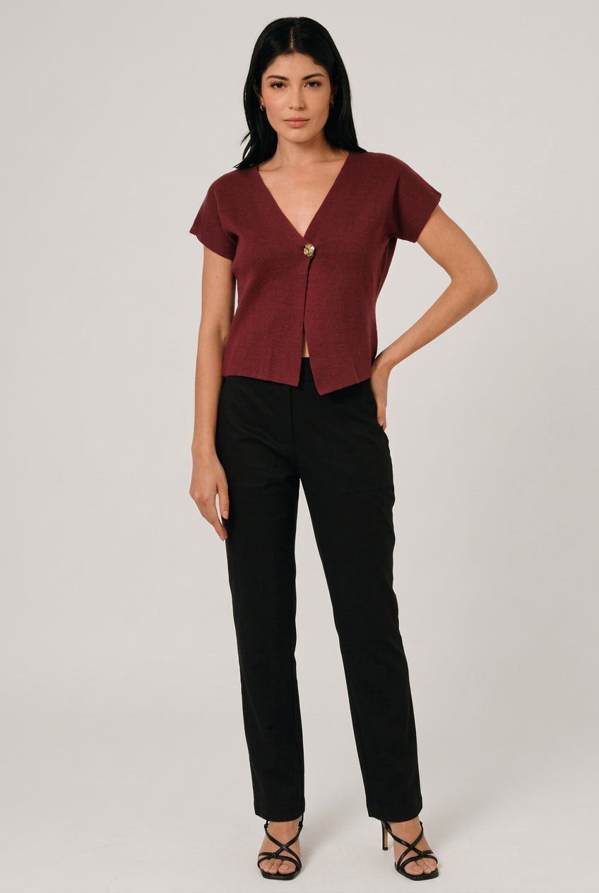 A woman wearing a short-sleeve burgundy knit top with a single decorative button, asymmetrical hemline and black pants, standing against a light curtain backdrop.