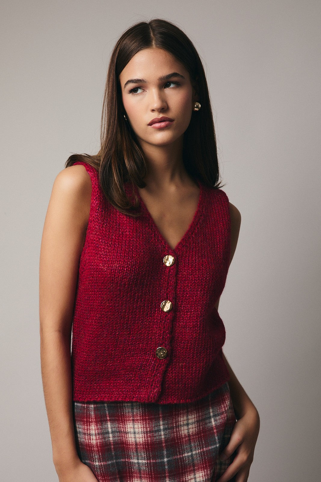 Woman wearing a red sweater vest over a plaid skirt against a plain background