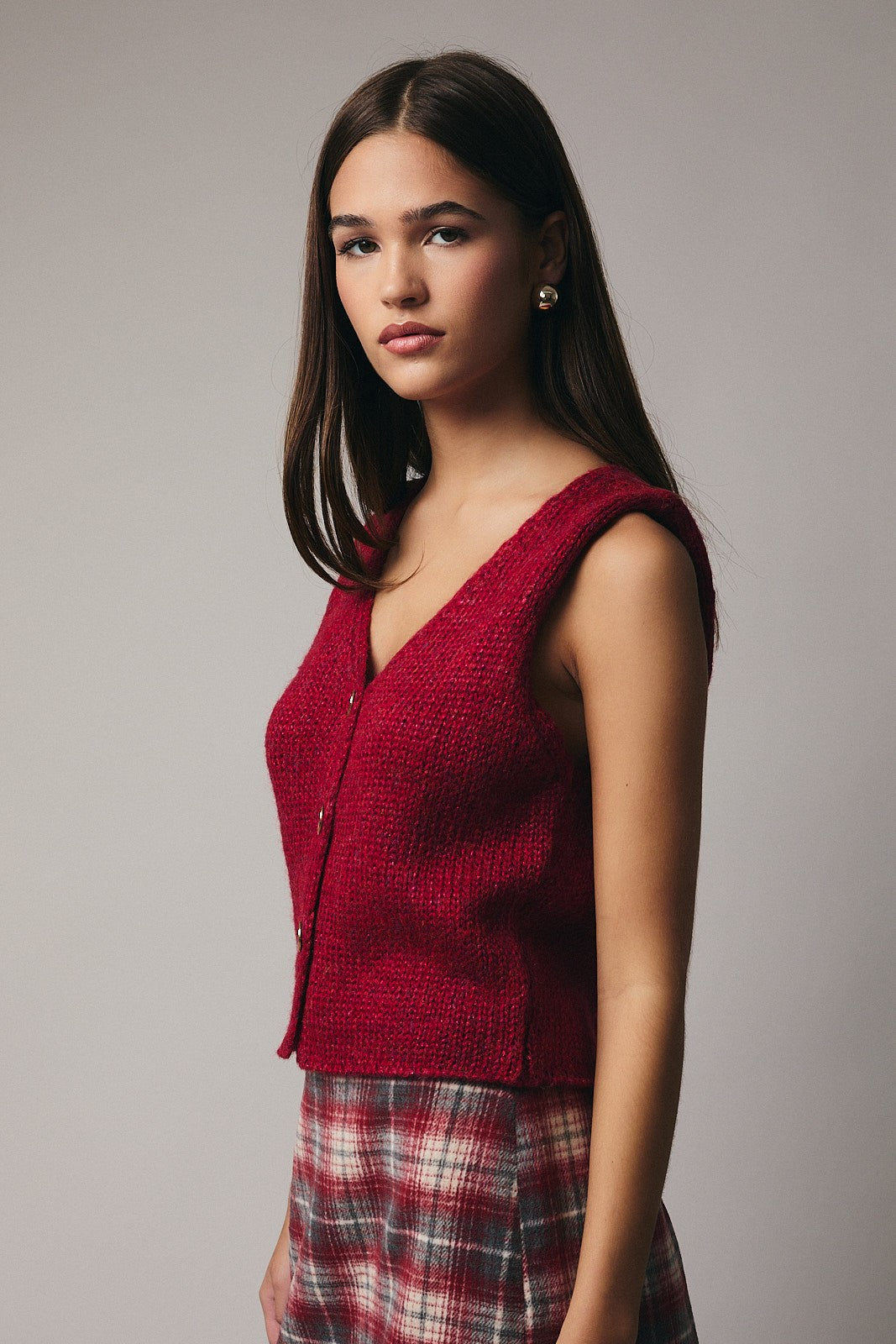 Woman wearing a red sweater vest over a plaid skirt against a plain background
