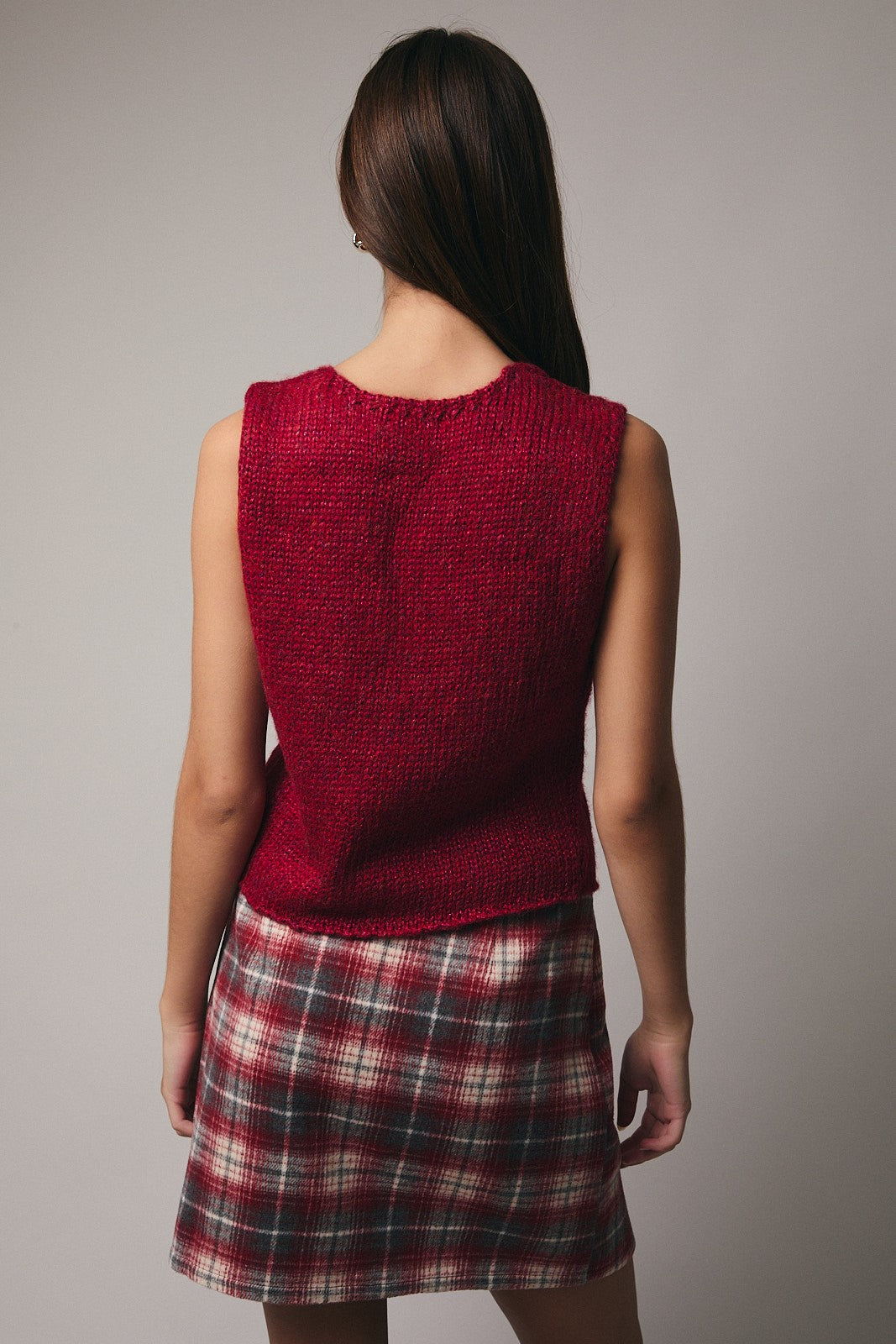 Back view of a woman wearing a red sweater vest over a plaid skirt against a plain background