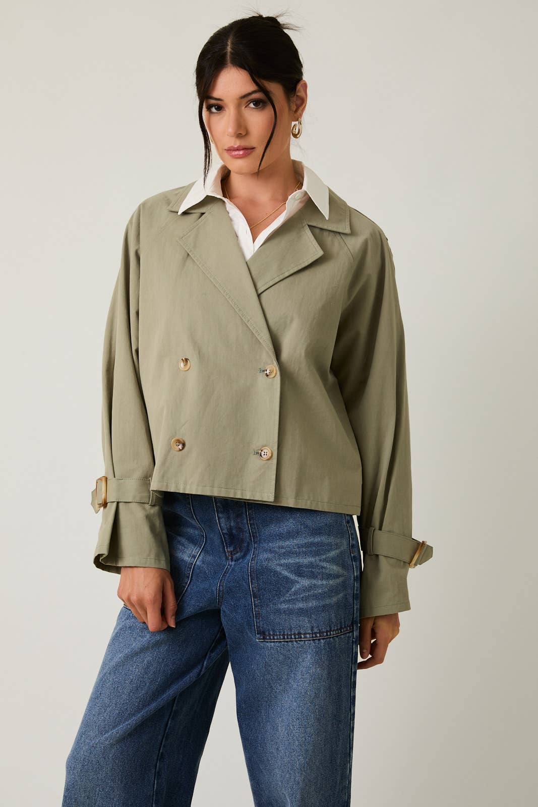 Woman wearing a sage green cropped trench jacket and blue jeans on a plain background
