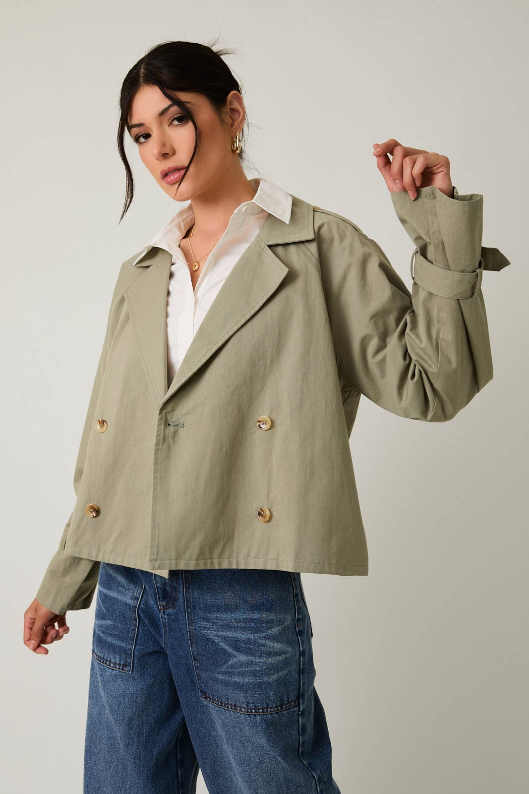 Woman wearing a sage green cropped trench jacket over a white shirt and blue jeans on a plain background