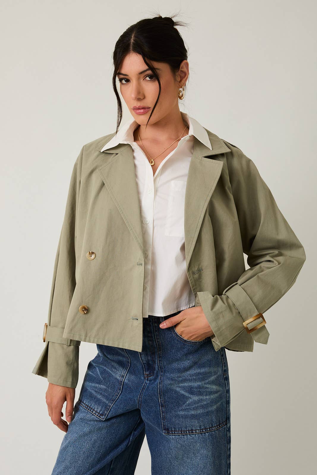 Woman wearing a sage green cropped trench jacket over a white shirt and blue jeans on a plain background