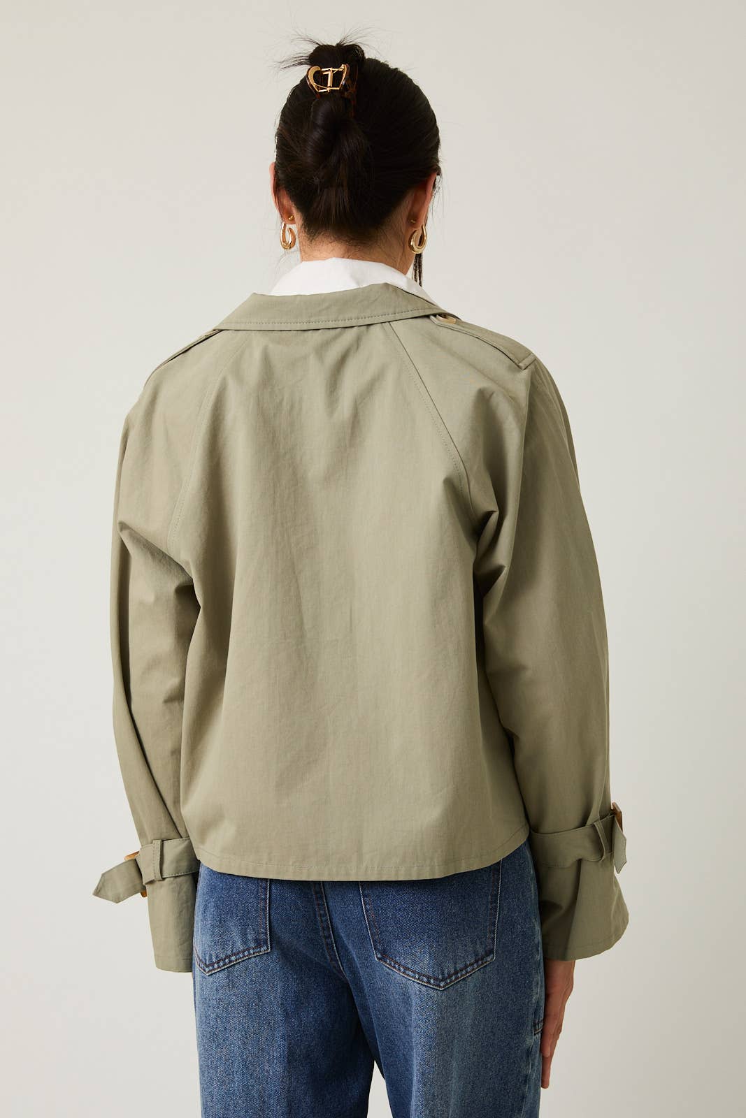 Woman wearing a sagae green cropped trench  jacket and blue jeans against a white background