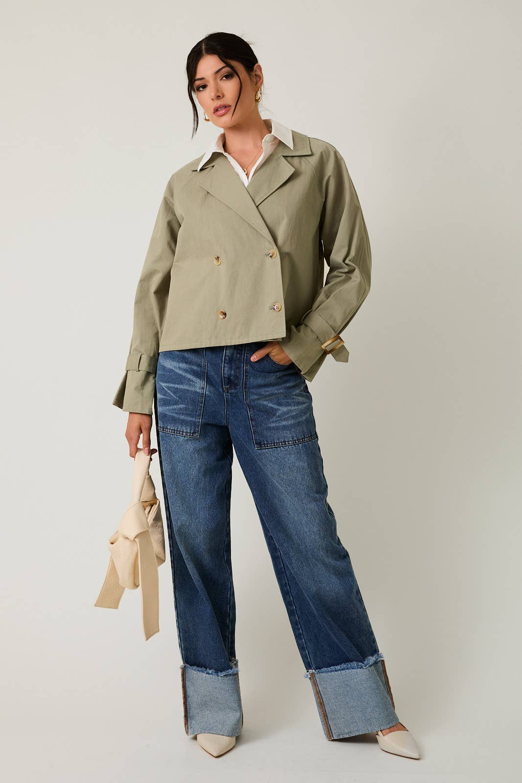 Woman wearing a sage green cropped trench jacket and blue jeans on a white background